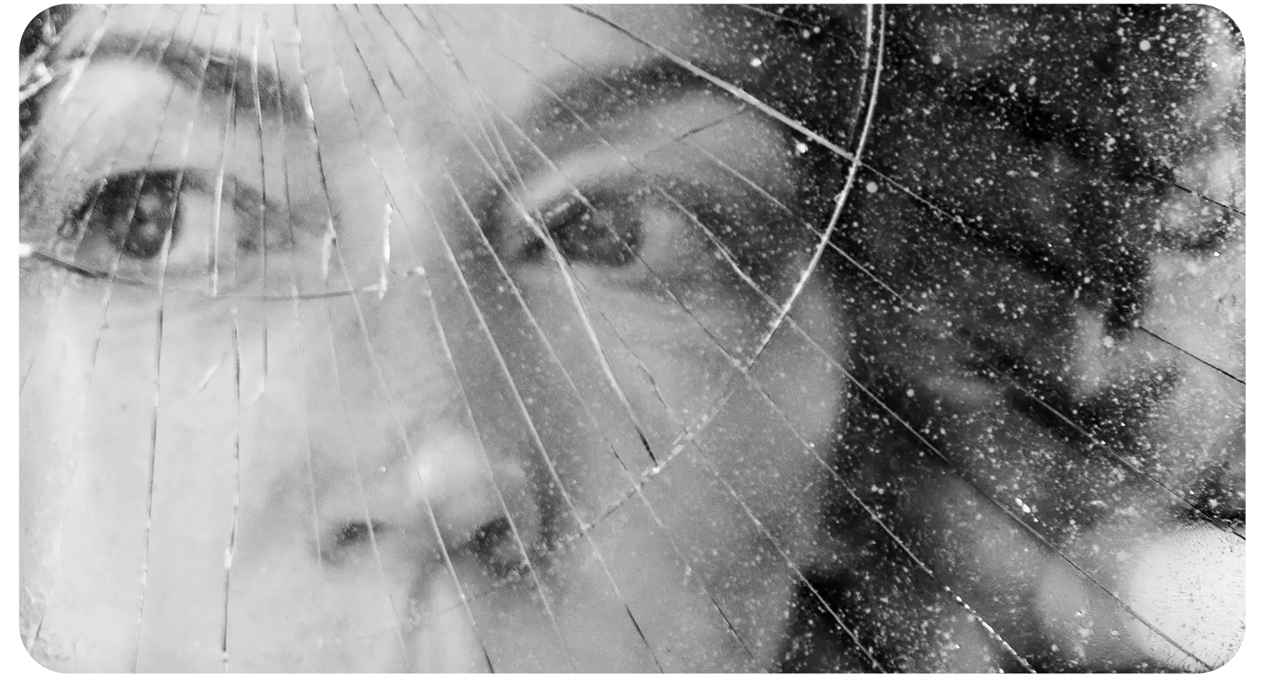 Black-and-white close-up of a person’s face seen through cracked, shattered glass, creating a fragmented and distressed visual effect